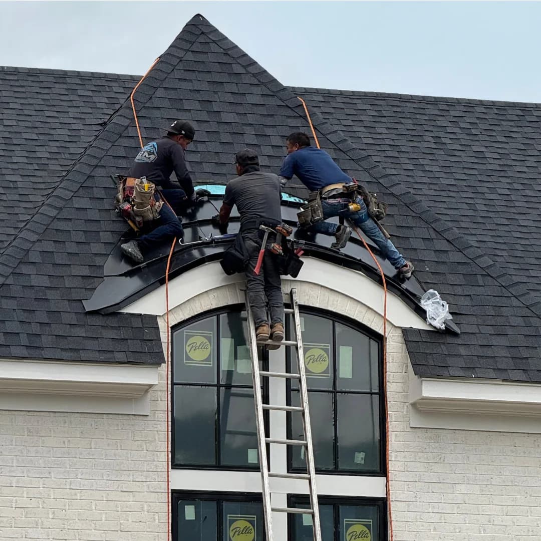 Three AEM roofers working at the ridge of a shingle roof above an arched window, ladder and safety lines set.