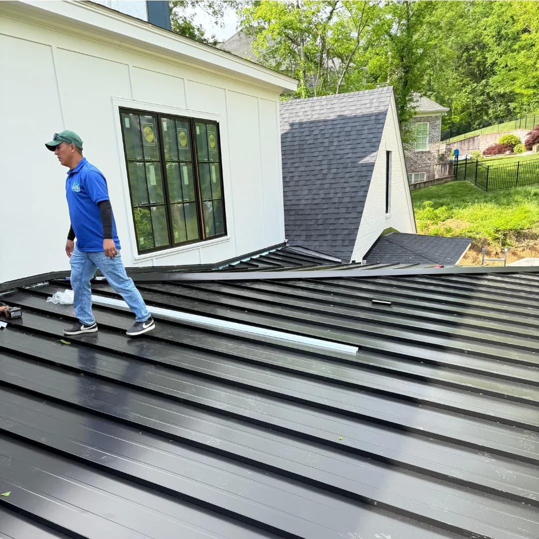 An AEM installer walking a run of dark standing-seam panels mid-install, the raised seams running downslope beside him.