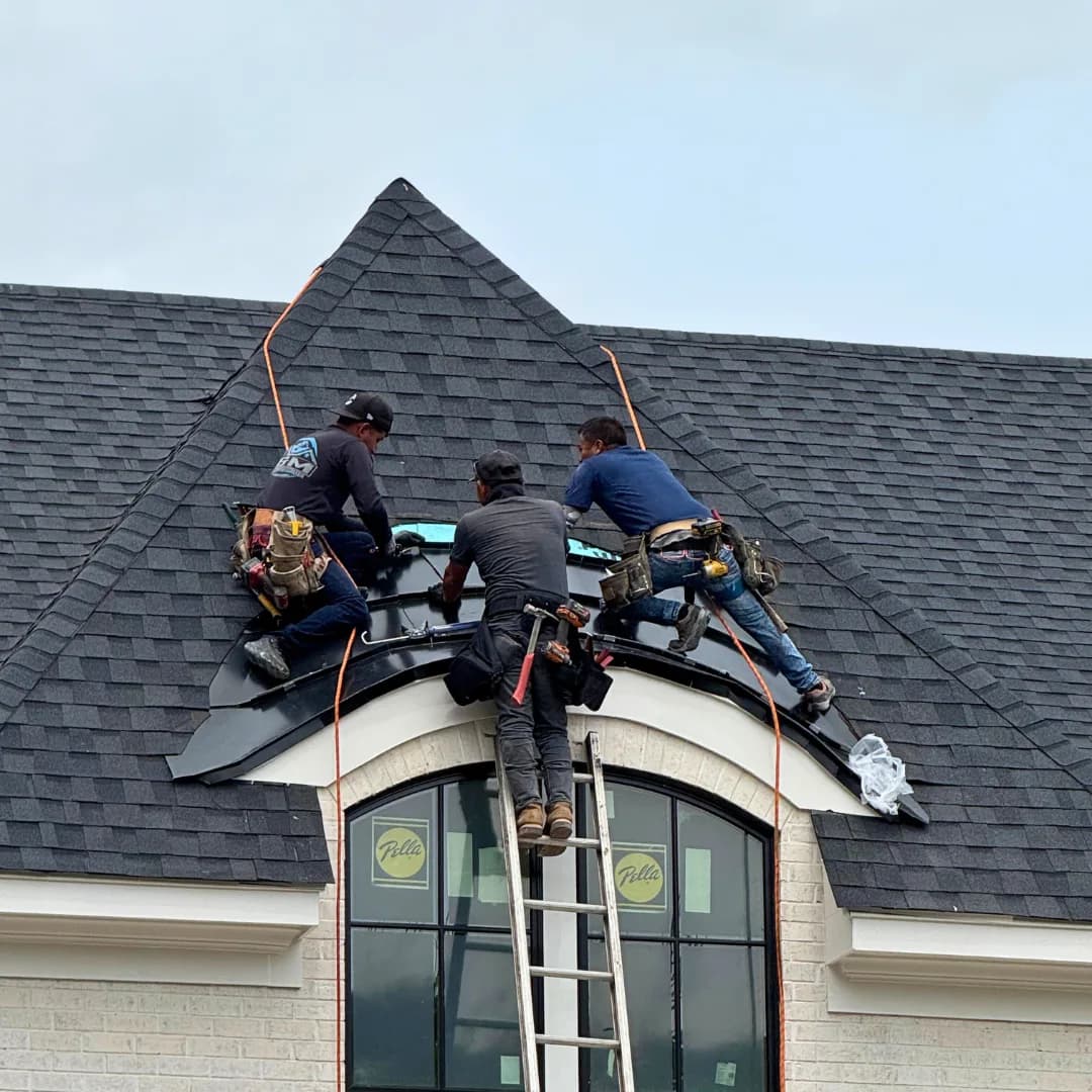 Three AEM crew members working from a ladder at the ridge of a shingle roof.