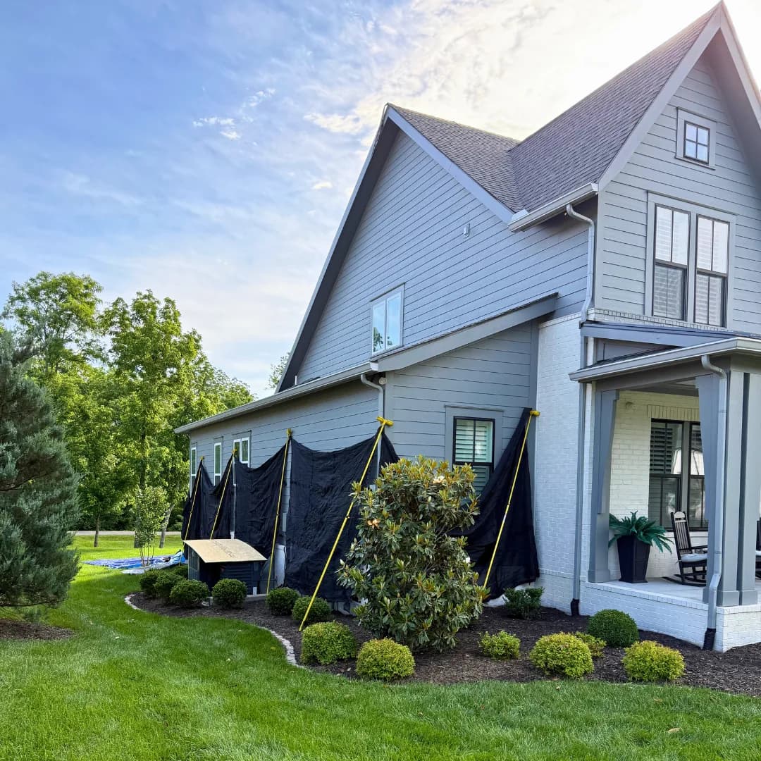 Gray farmhouse at golden hour with the roof replacement under way, black protection sheeting hung over the siding and the planting beds left untouched.