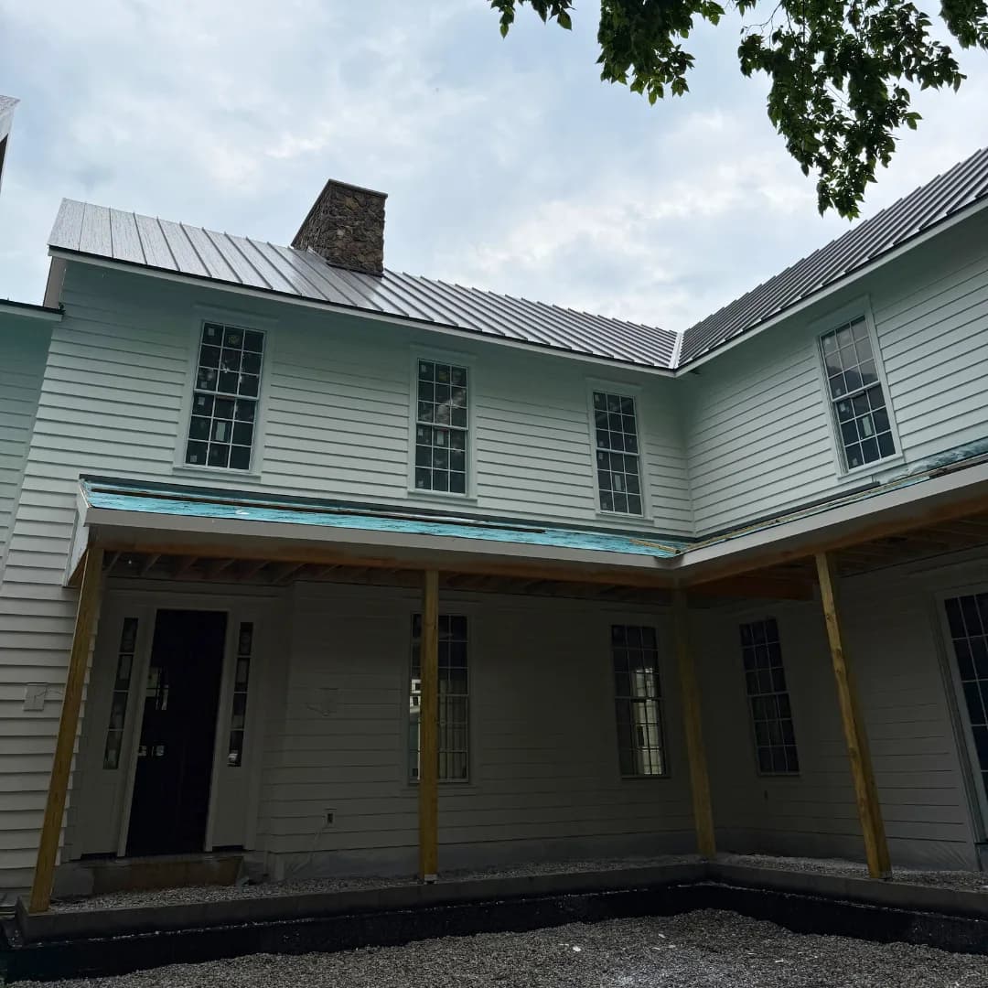 Standing-seam metal wrapping the inside corner of a clapboard farmhouse, stone chimney above the ridge.
