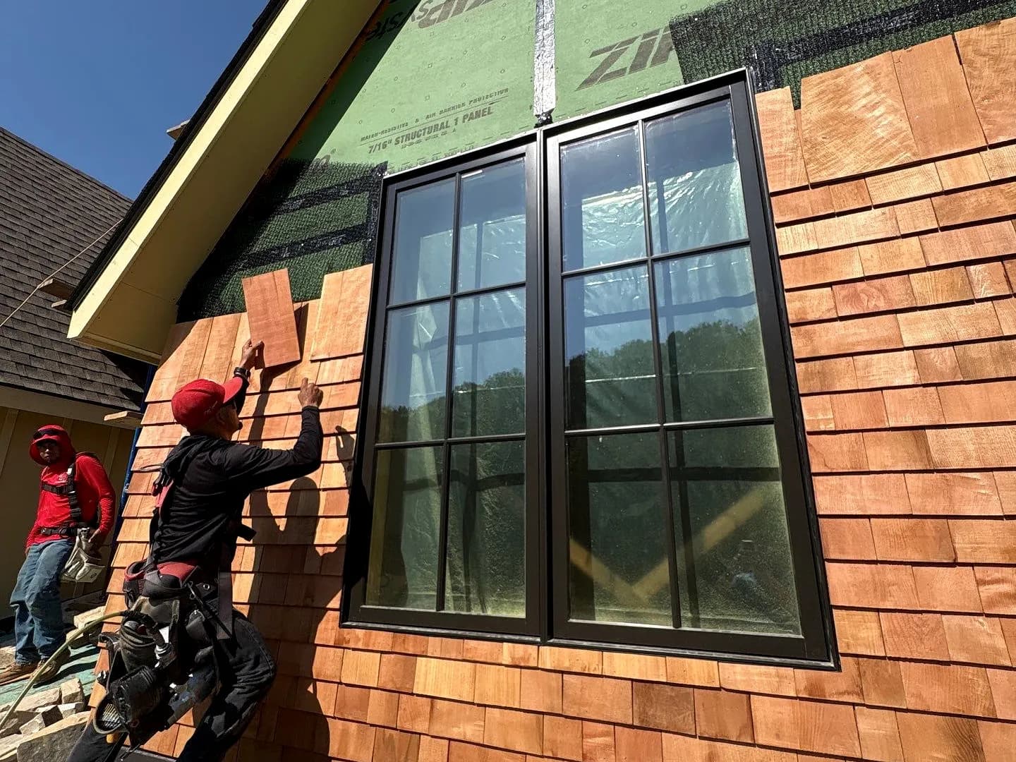 An AEM crew coursing cedar shake siding over ZIP System sheathing beside a tall window, the shakes running up to the gable.