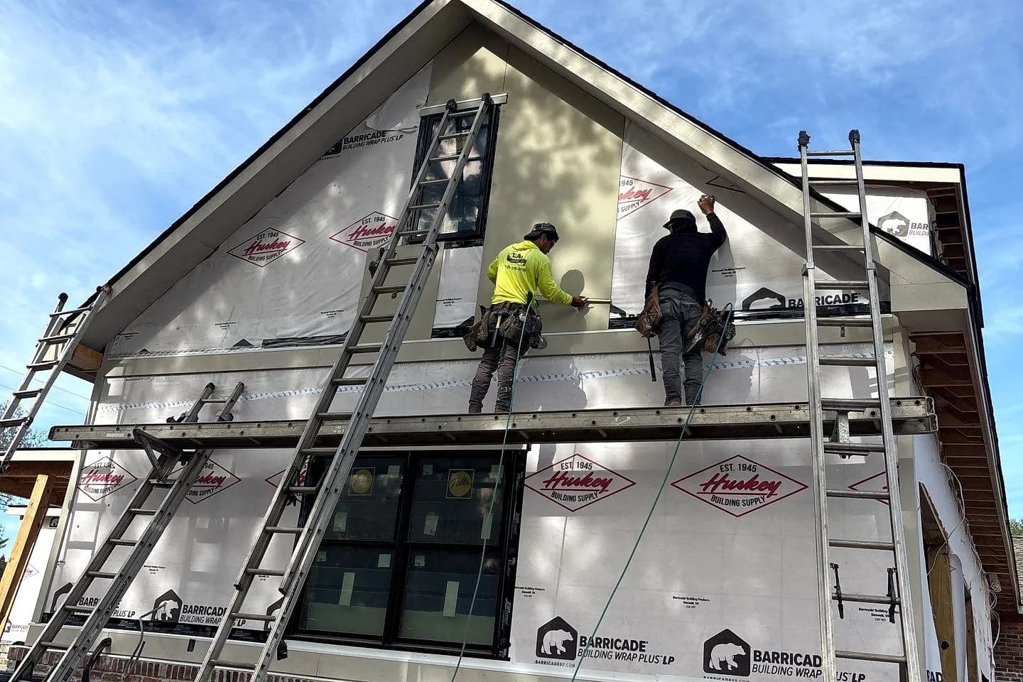 Two AEM crew working a wrapped elevation from a scaffold plank, the brick base below and the gable above still in building wrap.
