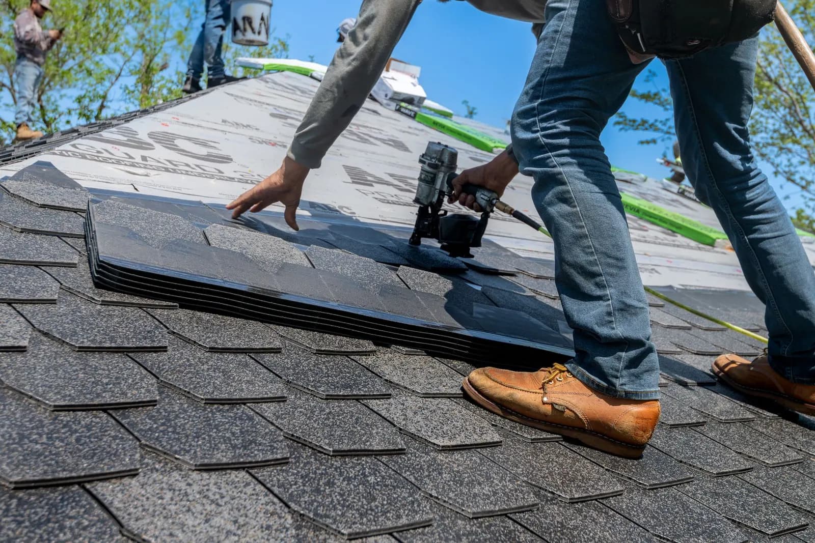 A roofer laying a course of asphalt shingles, nail gun in hand.